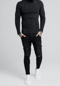 Schwarzer gestrickter Rollkragenpullover, eng anliegende, zerrissene schwarze Jeans und weiße Schnürsneaker, mit strukturiertem Stoff und verwaschenen Details.