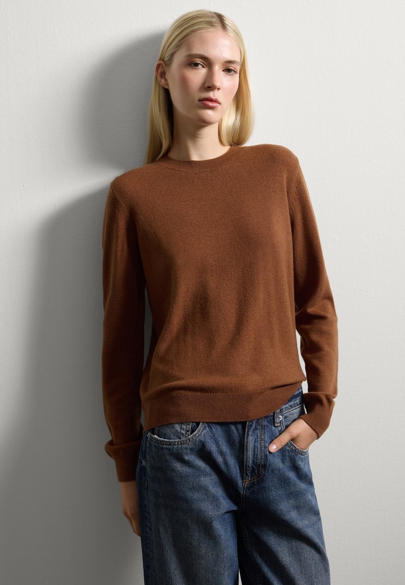 Junge Frau mit langen blonden Haaren, die einen braunen Pullover und eine blaue Jeans trägt, steht mit einer Hand in der Tasche vor einer schlichten Wand.