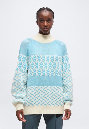 Jeune personne portant un pull tricoté à motif bleu clair et blanc avec des manches longues et un col haut crème, debout devant un fond uni.