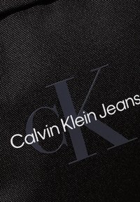 Sort tekstil taske med logoen "Calvin Klein Jeans" i hvidt og gråt skrift, med en glat, tekstureret overflade.