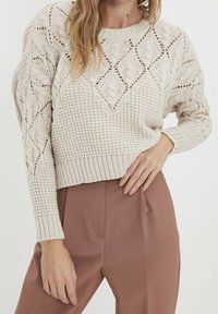 Světle béžový pletený svetr s diamantovými a texturovanými vzory, s žebrovanými manžetami a lemem, doplněný vysokým pasem růžových kalhot.