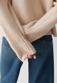 Mani incrociate dietro la schiena, indossando un maglione beige a costine e jeans denim blu, su uno sfondo neutro.