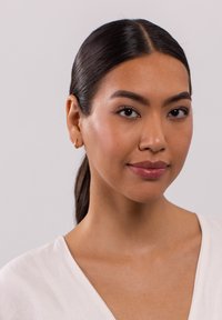 Goldene Creole im linken Ohr, glatte Haut mit natürlichem Make-up, schwarzes Haar zu einem eleganten Pferdeschwanz gebunden, trägt eine weiße Bluse mit V-Ausschnitt.