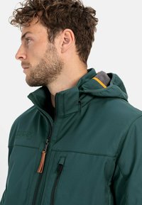 Grønne lynlåsejakke med en tekstureret overflade, der har en hætte og orange detaljer. Inkluderer kontrastfarvet foer og lynlåslommer.