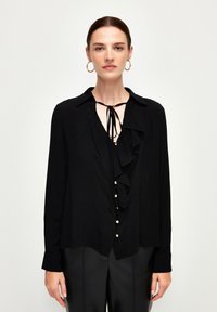Blusa nera a maniche lunghe con fronte arricciato, scollatura annodata, tessuto testurizzato e dettagli con bottoni, abbinata a pantaloni aderenti.