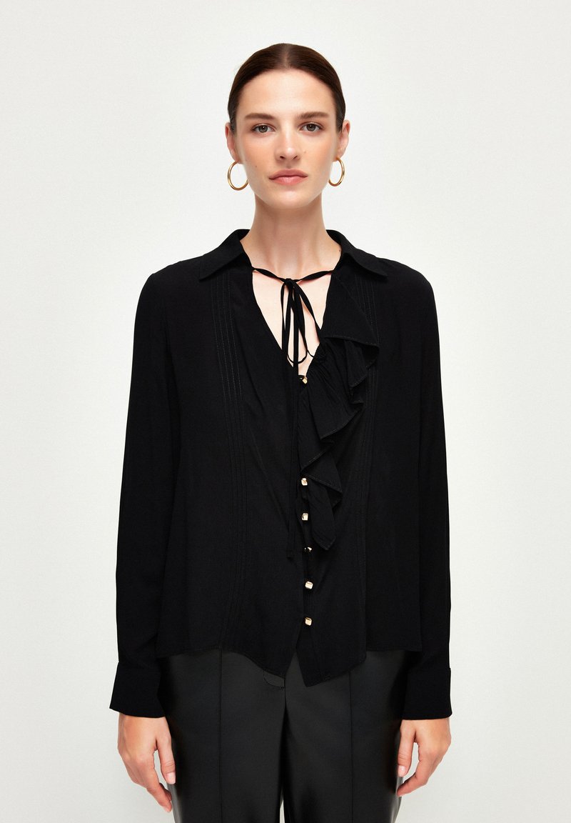 Blusa nera a maniche lunghe con fronte arricciato, scollatura annodata, tessuto testurizzato e dettagli con bottoni, abbinata a pantaloni aderenti.