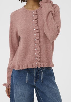 Vrouw draagt een stoffige roze gebreide cardigan met knopen en gerimpelde randen, gecombineerd met losse blauwe spijkerbroeken, staand tegen een effen achtergrond.