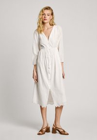 Vit medellång klänning med trekvartslånga ärmar, knappdetalj, vågig nederkant och broderat mönster. Stylad med bruna klacksandaler.