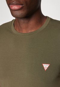 Nærbillede af en person iført en olivengrøn T-shirt med et lille rødt og hvidt Guess-logo på venstre bryst.