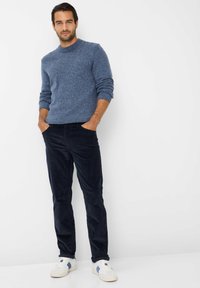 Blauwe gestructureerde gebreide trui, marinekleurige corduroy broek en witte sneakers met blauwe accenten, staand met handen in de zakken tegen een witte achtergrond.