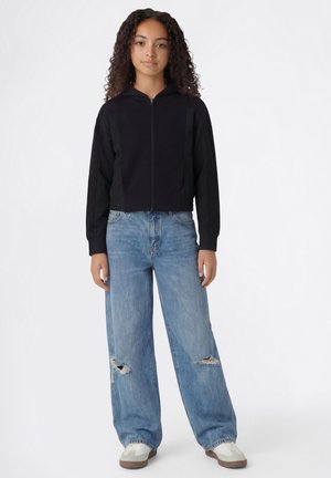 Jong persoon met krullend haar, gekleed in een zwarte hoodie met ritssluiting, gescheurde lichtblauwe wijd uitlopende jeans en witte sneakers, staande tegen een effen achtergrond.