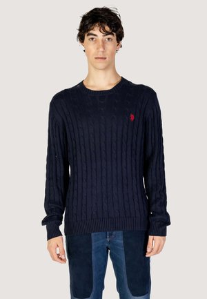 Maglione blu navy a coste con scollatura rotonda, maniche lunghe e un piccolo logo rosso sul lato sinistro del petto. Polsini e orlo a coste. Texture morbida.