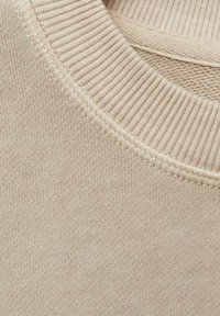 Beigem gestricktem Stoff, der eine Nahaufnahme des gerippten Ausschnitts und der glatten Textur zeigt, mit einem subtilen, einheitlichen Muster überall.