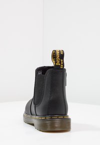 Black Chelsea boot with elastic side panels, yellow accent on heel tab, textured leather, and a brown, ribbed sole.