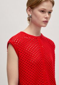 Rotes gestricktes Oberteil mit einem hexagonalen Durchbruchmuster, kurzen Ärmeln und einer Knopfleiste. Glatte Textur und lässiges Design.
