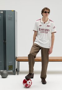 Camisola branca de manga curta com detalhes vermelhos e logótipo, combinada com calças castanhas claras e sapatos escuros, em pé sobre uma bola de futebol com padrão vermelho e branco.