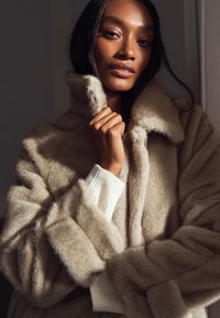 Beige jas van nepbont met een hoge kraag en een zachte textuur, gedragen over een wit geribd truitje. Voorzien van wijde mouwen en een losse pasvorm.