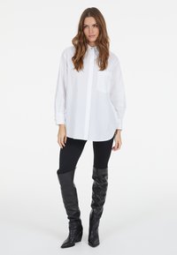 Weißes übergroßes Hemd mit Knöpfen und einer Brusttasche, kombiniert mit schwarzen Leggings und knielangen schwarzen Stiefeln. Flacher Kragen, lange Ärmel.
