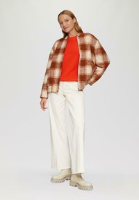 Kariertes Jackett in Braun und Creme, roter gerippter Pullover, weiße weite Hosen und beigen derben Stiefeln. Weiche Texturen und lässige Passform.