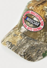 Casquette de baseball camouflage avec une visière incurvée, arborant un logo "NASCAR" brodé et un texte rouge sur un patch circulaire. La texture varie selon le tissu.