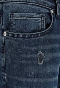 Tamno plave traperice s džepom koji ima vidljiv uzorak šavova i s istrošenim detaljima s poderanim rubovima.