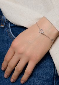 Srebrna bransoletka łańcuszkowa z wiszącym wisiorkiem w kształcie serca ozdobionym przezroczystymi kamieniami. Dłoń odpoczywająca na dżinsowej tkaninie, eksponująca wzór bransoletki.