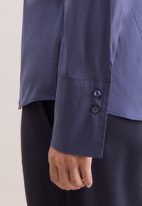 Donkerblauw shirt met lange mouwen en een gladde textuur. Heeft een manchetknoop met twee knopen en een nette zoom met subtiele stiksel details.