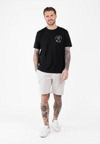 Mężczyzna z tatuażami, ubrany w czarną koszulkę z graficznym motywem palmy, beżowe szorty i białe trampki, stojący z rękami w kieszeniach.