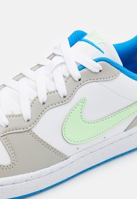 White and grey sneaker with light green swoosh, blue accents, and white laces. Features textured upper and rubber sole.