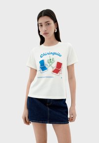 White cotton t-shirt featuring a colorful graphic of blue and red chairs, drinks, and "Chiringuito" text. Relaxed fit with short sleeves.