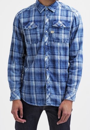 Uomo che indossa una camicia a quadri blu e bianca con tasche sul petto e pantaloni scuri, in piedi su uno sfondo bianco.