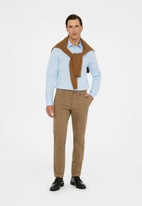 Camisa azul clara, suéter marrón colgado alrededor del cuello, pantalones de cuadros marrones y zapatos negros. Tela suave, corte a medida y estilo casual.