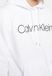 Sudadera con capucha blanca y un gran logo negro de "Calvin Klein", con cordones y un bolsillo frontal. Textura de tela suave.