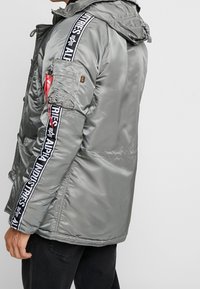 Grå vadderad jacka med huva, med glansig yta, svarta och vita logoband på ärmarna, och en ficka med dragkedja på armen.