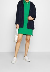 Vihreä polo-mekko, tummansininen corduroy-takki, valkoiset lenkkarit ja pieni valkoinen olkalaukku. Suuret ja sileät pinnat sekä rento muotoilu.