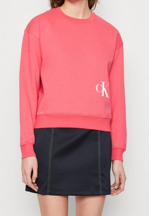 Personne portant un sweat-shirt corail à manches longues avec un logo « cK » blanc et une minijupe noire avec des coutures verticales blanches.