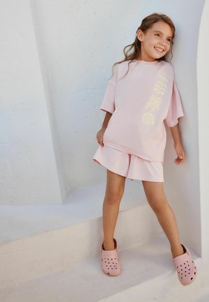 Smiling girl in light pink oversized T-shirt with patterns, matching shorts, and pink slip-on shoes, standing against white textured wall.