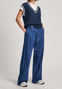 Osoba v bílém tričku s námořnickým modrým pleteným vestou, širokých modrých džínech s rukama v kapsách a bílých teniskách stojící na světlém pozadí.