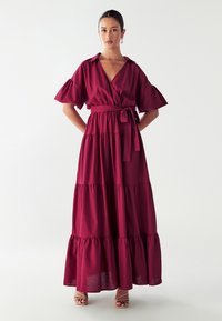 Wickelstil, kurzärmliges Maxikleid in tiefem Burgunderrot, mit gebundenem Taillengürtel und gestuften Rock mit Rüschenakzenten. Glatte Stofftextur.