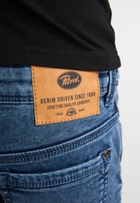 Blå cowboybukser i denim med en brun læderlap mærket "Petrol", med syningsdetaljer og en klassisk pasform. Sort skjorte synlig ovenover.
