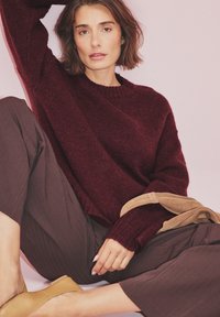 Burgundowa sweter z dzianiny z okrągłym dekoltem, w połączeniu z luźnymi brązowymi spodniami. Wyraziste beżowe akcesoria i żółte mokasyny.