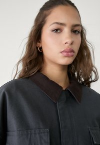 Dunkelgraue Jacke mit einem braunen Cordkragen, ausgestattet mit zwei Fronttaschen und einer lässigen Passform. Glatte Stofftextur mit minimalen Details.