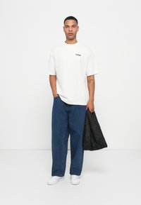 Jonge man in een wit hummel t-shirt, blauwe wijde jeans, witte sneakers, die een zwart gevoerd jack vasthoudt in een witte studio.
