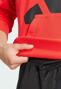 Felpa rossa con un design nero, caratterizzata da un orlo a coste. Una mano afferra l'orlo, rivelando il suo tessuto testurizzato. Indossata con pantaloni corti neri con vita elasticizzata.