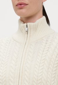 Κρεμ πουλόβερ με καλωδιωτό σχέδιο και ψηλό γιακά, διαθέτει μπροστινό φερμουάρ και ριγωτή υφή, προσδιορίζοντας ζεστασιά και άνεση.