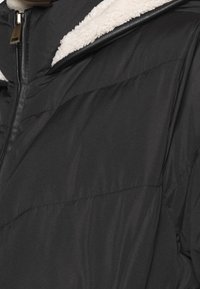 Veste matelassée noire avec une capuche douce doublée en polaire. Dispose d'une fermeture éclair à l'avant et d'une texture matelassée avec des coutures horizontales.