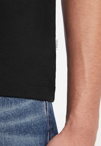 Melnas kokvilnas T-krekls ar taisnu apakšmalu, uz kura sānu ir mazs balts "GUESS" apzīmējums, valkāts kopā ar gaiši ziliem džinsiem.