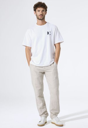 Camiseta blanca de algodón con un pequeño logo oscuro en el pecho, combinada con pantalones de lino beige claro y zapatillas blancas. Corte sencillo y relajado.