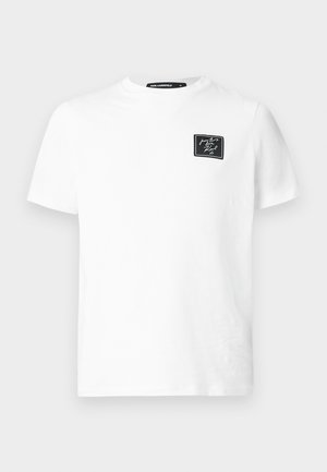 Biała koszulka typu t-shirt z krótkim rękawem i okrągłym dekoltem, z małą czarną prostokątną naszywką na lewej piersi z białym tekstem pisanym kursywą.