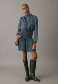 Vestido tipo smock en tejido azul texturizado con manga larga, escote en forma de ojo de cerradura, cintura ajustada y dobladillo con volantes en capas; combinado con botas negras hasta la rodilla.